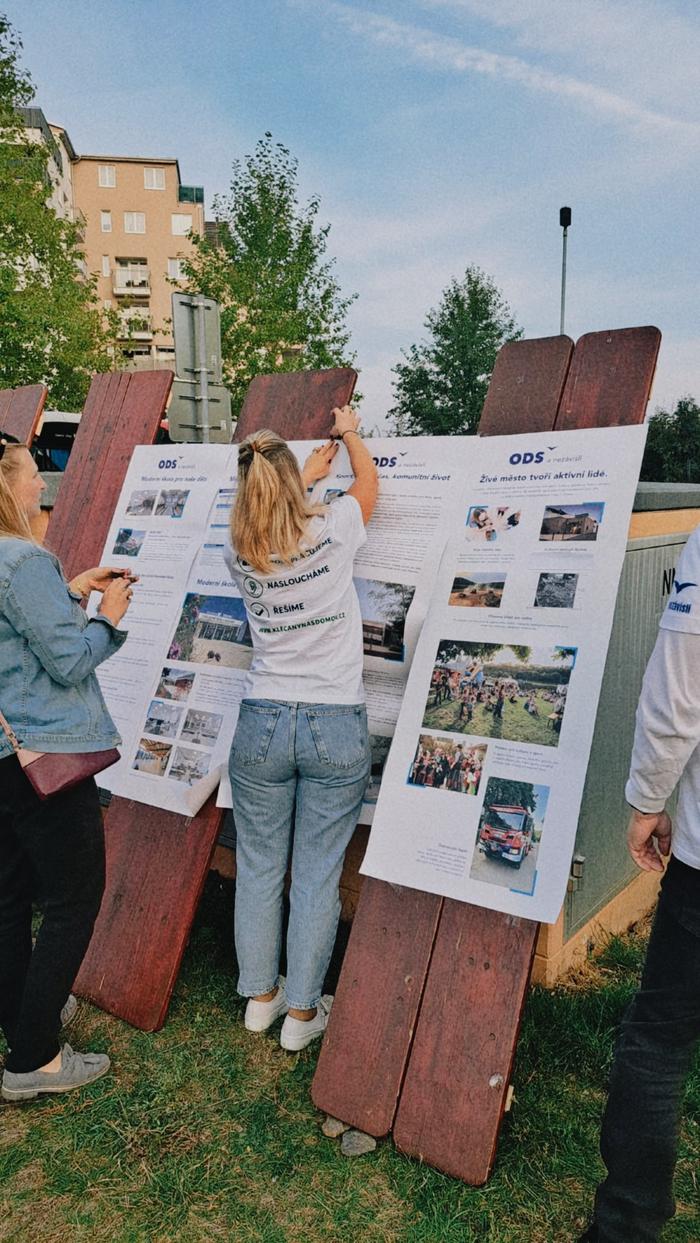 Žena v tričku s nápisem Jdeme za vámi připevňuje plakát na informační panel
