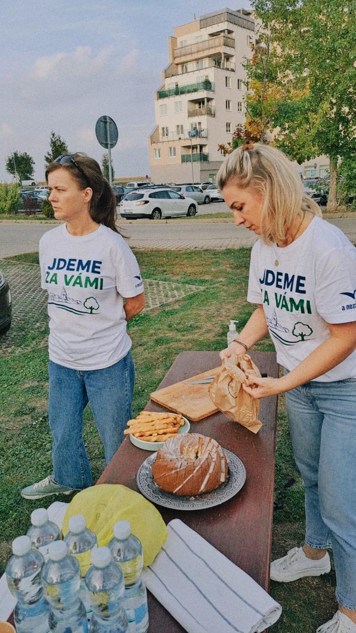 Dvě ženy v tričkách s nápisem Jdeme za vámi chystají na stůl bábovku a pečivo