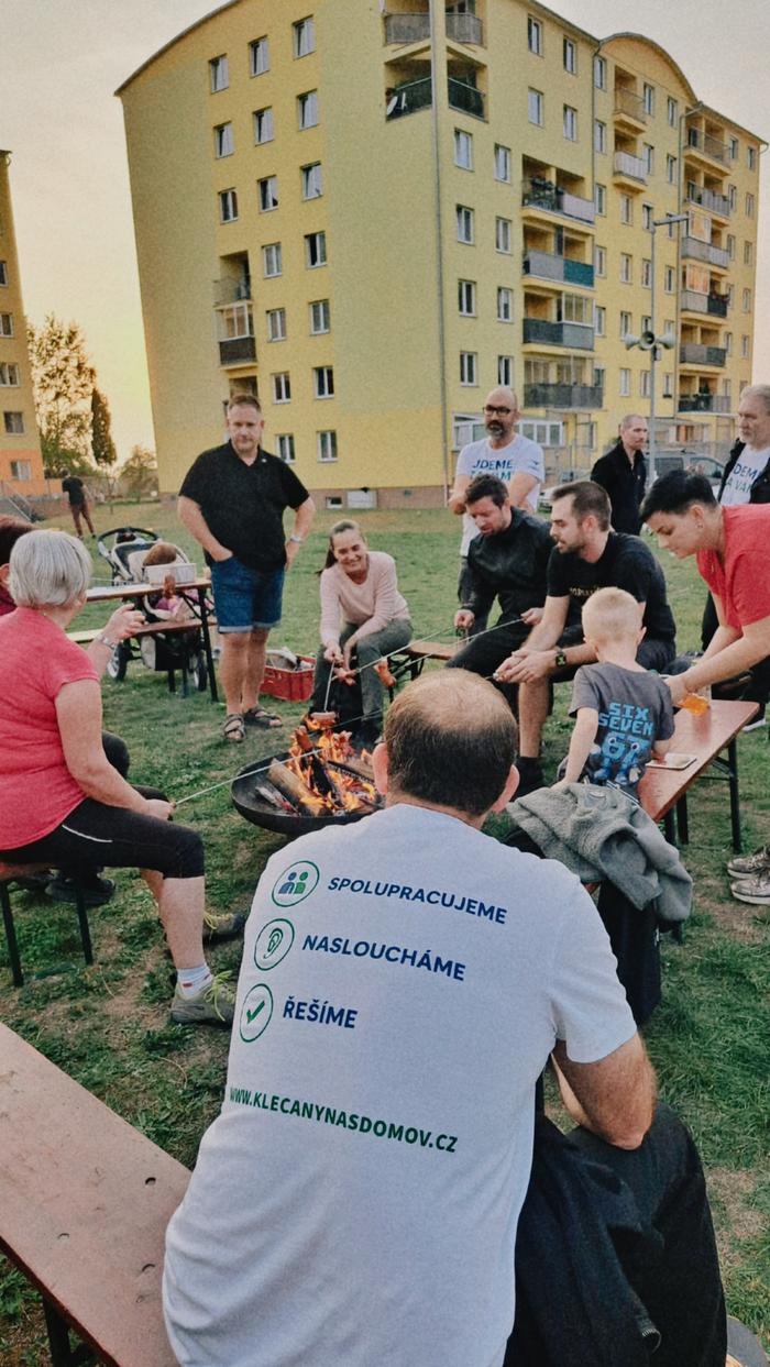 Lidé opékají u ohniště, v popředí muž v tričku s nápisy Spolupracujeme, Nasloucháme, Řešíme