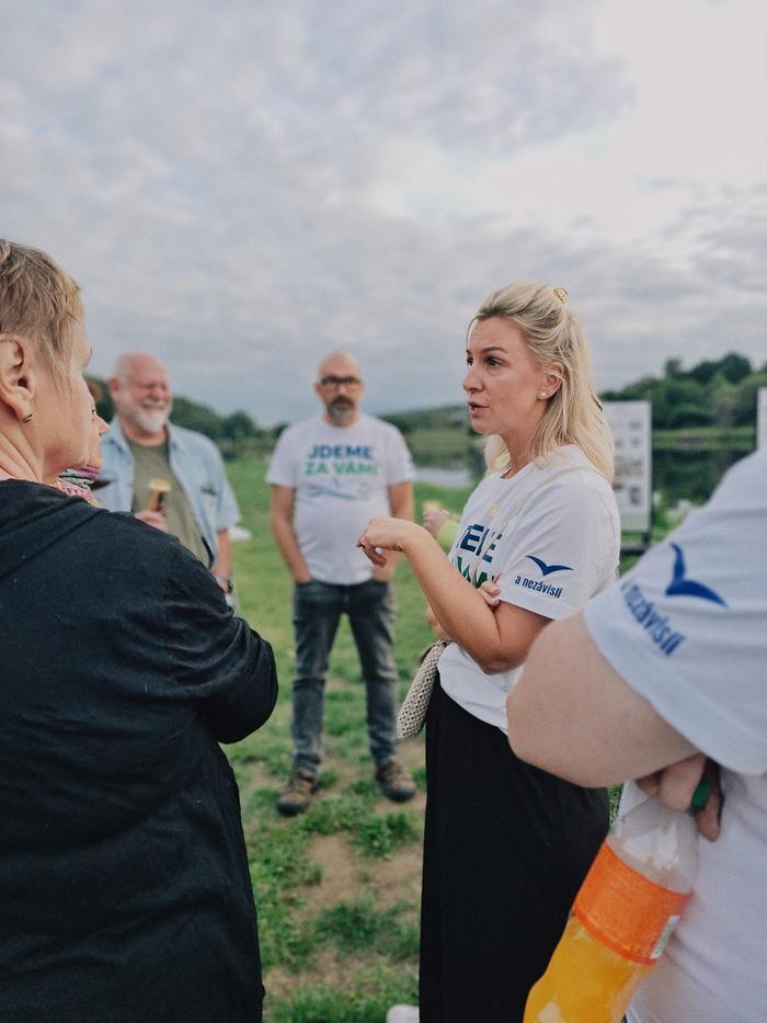 Žena v tričku s nápisem Jdeme za vámi mluví s návštěvníkem, kolem stojí další účastníci
